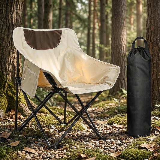 A folding camp chair with a cream and brown fabric seat, shown outdoors in a forest beside a black drawstring storage bag.